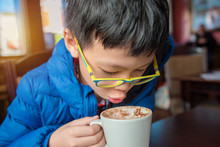 Young Asian Boy Blowing Hot Chocolate Drink In Cafe