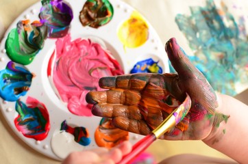 Wall Mural - Child painting her hand with a paintbrush and background with palette of colorful paint