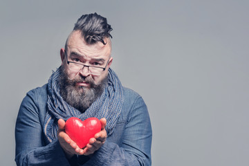 Requesting bearded man in a blue jacket and scarf holding red heart. Gray background