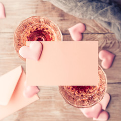 Two stemmed champagne glasses with pink hearts blank paper note card to fill with your text on wooden textured background. Valentine's day wedding romantic date invitation. Toned