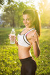 © Bojan - Young smiling woman holding bottle of water and showing thumb up outdoors.