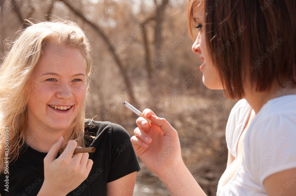 Two girl friends smoking outside next to a river in the woods. Teenage  girls sneak a cigarette outside away from parents. Teenagers being  mischievous. Stock-foto