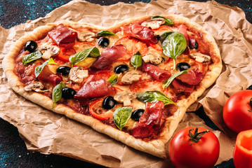 Heart shaped pizza with tomatoes and prosciutto for Valentines Day on vintage black turquoise background. Food concept of romantic love