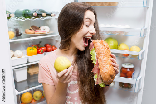 Fotografía Young woman choosing between apple and big sandwich standing in front of the ref