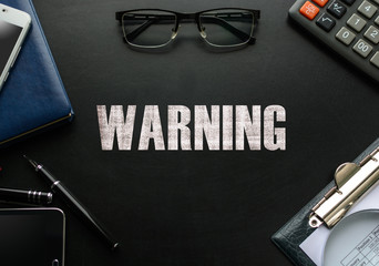 Black chalkboard with business accessories (notepad, magnifying glass, fountain pen, tablet, phone, glasses and calculator) and text WARNING. Top view.
