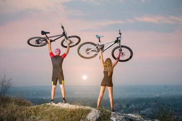 Man and woman bikers holding bikes high up in the sky on the top of a hill against magnificent sunset with blurred background. Pink Kinesio tape glued on the girl's hand.