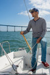 A man in a striped sweater, jeans, a blue cap and red sneakers controls sailing boat. Boy holds the tiller of a sailing boat in the hands. The bridge on background. Adventure. Sailor