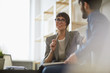 © pressmaster - Two cheerful colleagues smiling while discussing something in modern office, focus on stylish businesswoman wearing creative haircut and glasses
