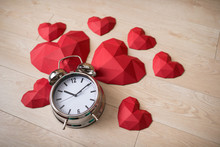 Closeup Of Big Alarm Clock With Many Red Polygonal Paper Heart Shapes Over Wooden Bachground