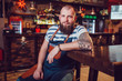 © kanashkin - Bearded barman with tattoos wearing an apron sitting at the bar.