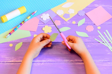 Wall Mural - Small child does a greeting card for mom. Child holds scissors in hand and cuts a leaf from paper. Colored paper, paper templates, glue stick, pencil on a table. Mother's day or March 8 easy crafts