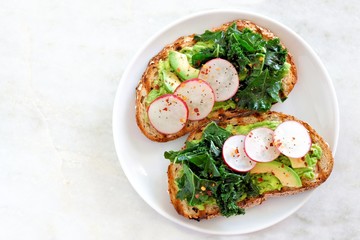 Wall Mural - Plate of avocado toast with kale and radish on whole grain bread, top view on a marble background