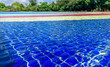 © shumoff - Light blue, red, yellow and blue striped mosaic floor of fountain covered with clear water at sunny day on the background of blurry green trees