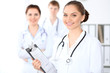 © rogerphoto - Happy female doctor keeping medical clipboard while medical staff are at the background. Successful team at health care and medicine concept