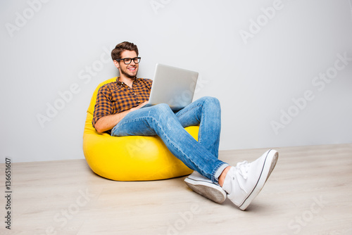 Fotografía Happy young man sitting in yellow pouf and using laptop