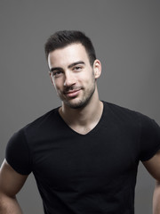 Wall Mural - Moody portrait of confident young smiling fitness male model in blank black shirt over gray studio background. 