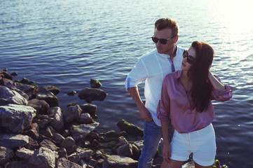 Young Happy fashionable couple resting by the sea on sunset. The man and woman Wearing in romantic sexy summer clothes and  in stylish trendy sunglasses