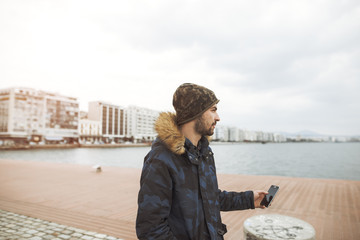 young man dressed in khaki fashion and a blue camo jacket is walking on the city dock while holding a phone