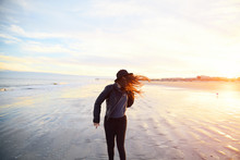 Young Woman With Long Curly Hair Shaking Head On Beach On Sunset. Girl In Black Hat Having Fun On Ocean Coast. Female In Leather Jacket Standing On Wet Sand. Calming Waves, Pastel Cloudy Sky.