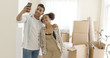 © Dash - Happy young couple posing for a selfie in their new home in front of a pile of brown cardboard boxes