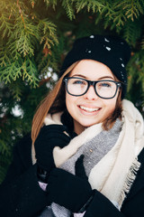 Wall Mural - Cheerful beautiful girl with blue eyes in black glasses in a black hat and coat with scarf in the winter park on the background of christmas tree