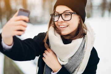 Wall Mural - Happy cheerful girl with blue eyes in black glasses in black coat and hat with scarf doing selfie on her smartphone