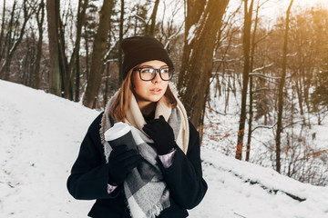 Wall Mural - Portrait of a beautiful girl in black glasses in a black hat and coat and gloves with scarf in the winter park holding warm coffee