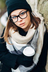 Wall Mural - Portrait of a beautiful girl with blue eyes in black glasses in a black hat and coat and gloves with scarf in the winter park holding warm coffee