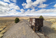 Old Mining Ore Cart On Tracks Underneath A Beautiful Blue Sky With Clouds In The Nevada Desert At The Berlin Ghost Town.