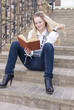 © danmorgan12 - Portrait of Cute and Tranquil Caucasian Blond Woman Reading Book While Sitting Straight on Stairs Outdoors.