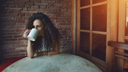 Portrait of young brunette female drinking tea and thoughtfully looking out of the coffee shop window while enjoying her leisure time alone, beautiful curly girl lunch in cafe during her work break