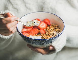 Healthy breakfast greek yogurt, granola and strawberry bowl in hands of woman wearing white loose knitted woolen sweater, selective focus. Clean eating, healthy, vegetarian, dieting food concept