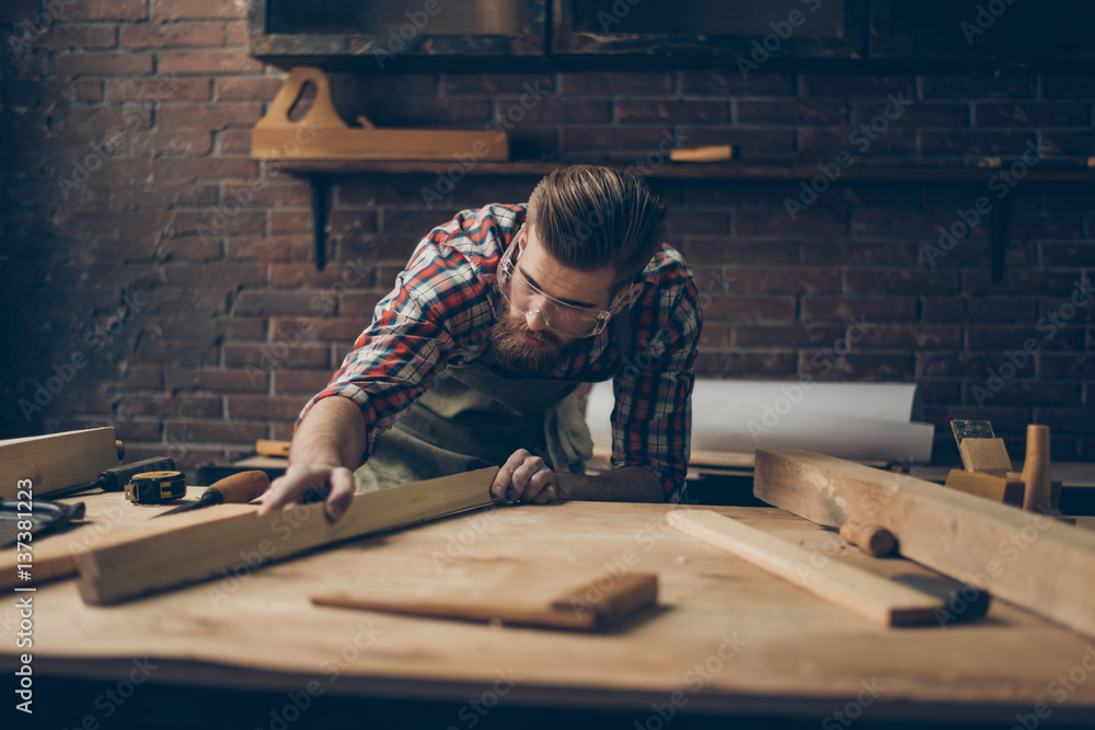 Bearded handsome cabinetmaker at the tabletop with tools.  Stylish craftsman with brutal hairstyle and saved glasses holding woodenplank at his workstation - obrazy, fototapety, plakaty 