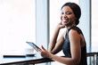 © nakophotography - Young well dressed black female holding a digital electronic tablet while smiling happily at the camera, while seated at a counter next to large bright windows.