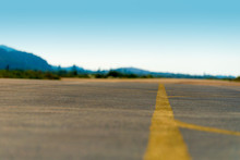 Selective Focus On An Empty Airfield Runway With Yellow Direction Lines. Blue Sky And Mountains In The Background.