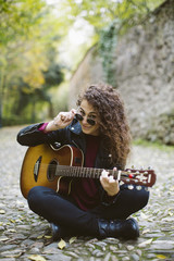 Beautiful young woman looking at the camera with a guitar on forest. Girl wearing black jacket and sunglasses, fashion lifestyle.