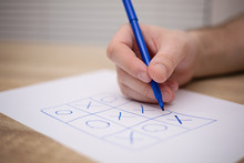 Male Adult Hand Holding Blue Pen Above A Paper With A Tic-tac-toe Game