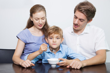 Wall Mural - Displeased little boy eating porridge with parents