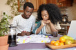 © wayhome.studio  - Young African couple doing paperwork together, sitting at kitchen table with lots of papers, calculator and laptop, looking frustrated. Dark-skinned family calculating domestic budget at home