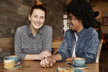 Happy lesbian couple having date at cafe. Cute Caucasian redhead girl holding hands with her dark-skinned female spouse while spending nice time together at restaurant, drinking coffee and eating cake