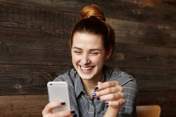 Cheerful young redhead female student with cute smile siting in modern cafe interior, using cell phone, checking newsfeed on her social network accounts. Pretty girl surfing internet on mobile