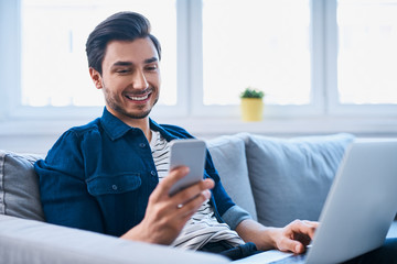 Young man sitting relaxed on sofa with laptop checking his smartphone messages