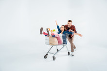 Happy Young Man Pushing Shopping Cart With Excited Woman And Grocery Bag On White