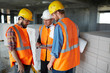 Group of three workmen wearing protective helmets and vests standing among concrete walls on construction site discussing development progress with foreman inspector