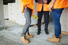 Low Section Shot Of Legs Of Two Construction Workers Wearing Jeans And Brown Leather Work Boots Standing With Man In Suit On Concrete Floor
