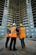 © pressmaster - Back view portrait of three workmen wearing reflective orange vests and hard hats standing on construction site against unfinished apartment building