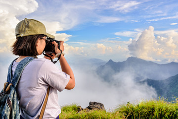 Hiker asian teens girl with digital camera and backpack taking photo beautiful natural of sierra sky clouds and fog during sunset in winter on mountain at Phu Chi Fa Forest Park, Chiang Rai, Thailand