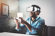© SFIO CRACHO - Concept of technology,gaming,entertainment and people.African man enjoying virtual reality glasses while relaxing in living room.Happy young guy with VR headset playing video game at home.Blurred.