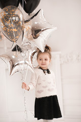 Poster - Smiling baby girl 5-6 year old holding silver star balloons in white room. Wearing knitted sweater with hearts and black dress. Birthday party. Celebration.