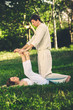 Young man and woman dressed in a white robe doing thai massage with yoga exercises lying on the mat. Sunny summer park with green lawn in the background